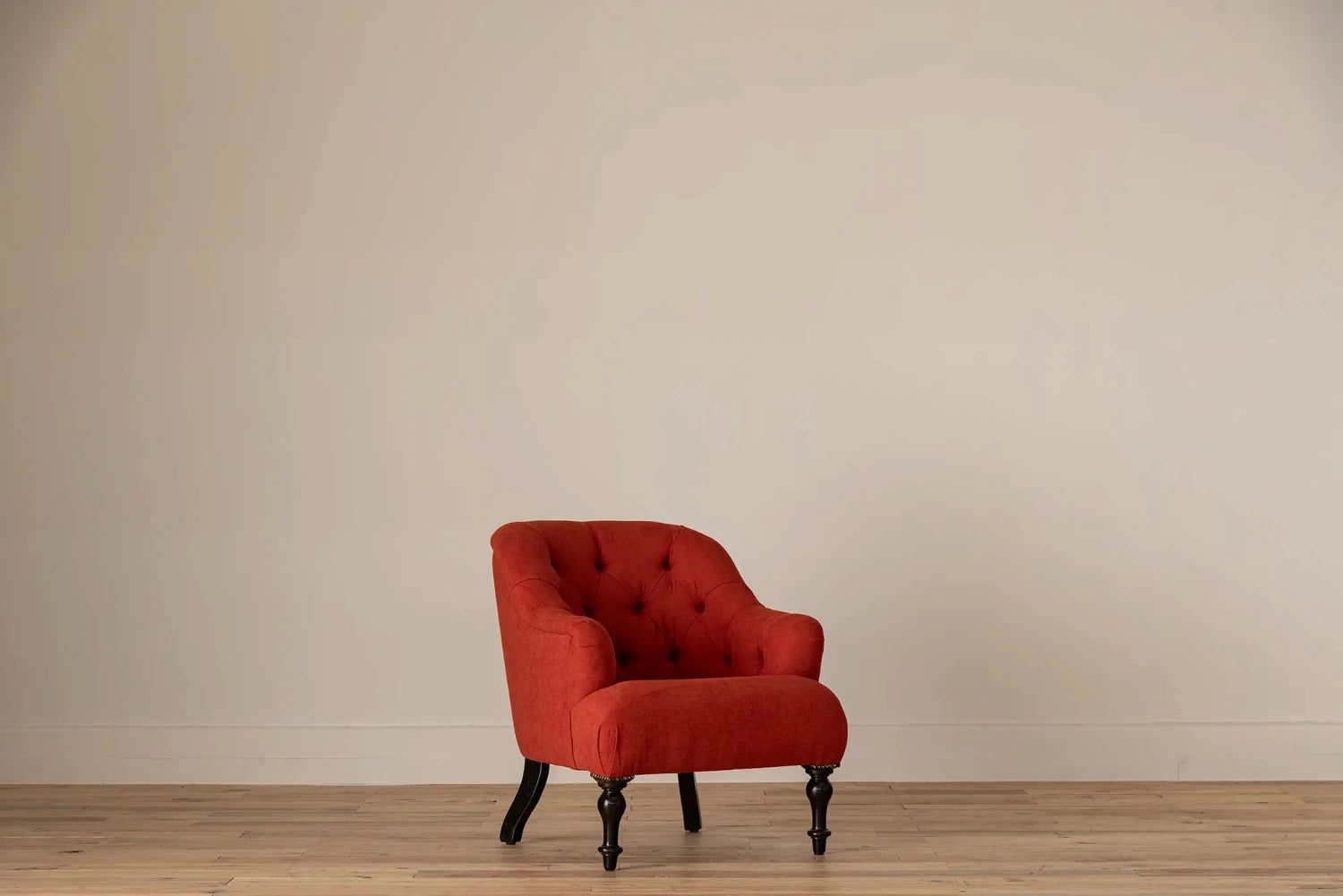 Rust-red button-tufted armchair with nailhead trim and turned dark wood legs, set against a cream wall and light wood floor