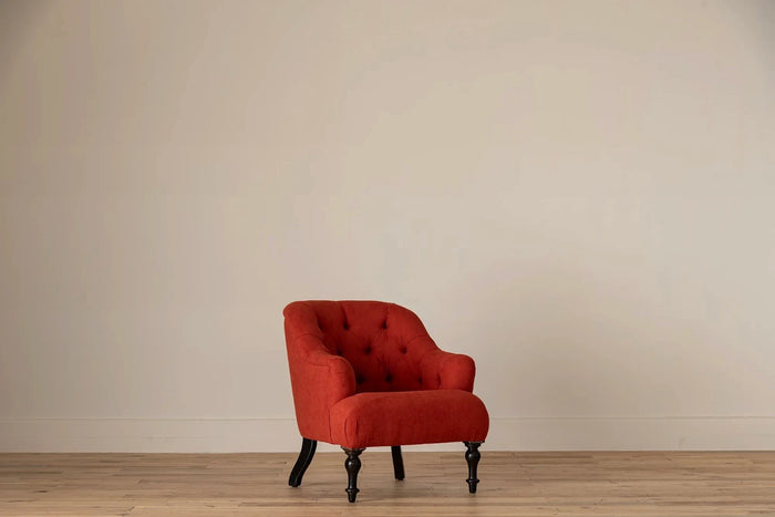 Rust-red button-tufted armchair with nailhead trim and turned dark wood legs, set against a cream wall and light wood floor
