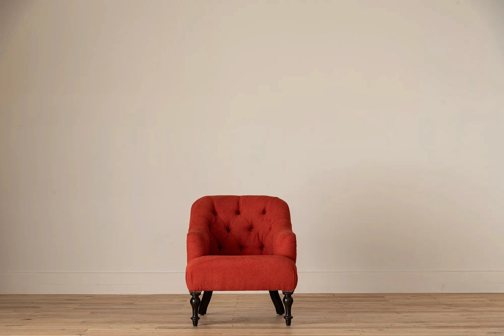 Rust-red button-tufted armchair with nailhead trim and turned dark wood legs, set against a cream wall and light wood floor