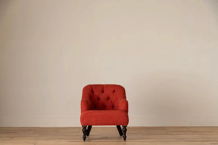 Rust-red button-tufted armchair with nailhead trim and turned dark wood legs, set against a cream wall and light wood floor