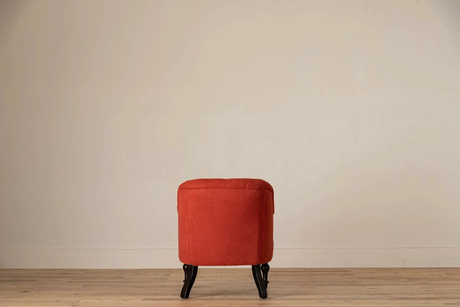Rust-red button-tufted armchair with nailhead trim and turned dark wood legs, set against a cream wall and light wood floor