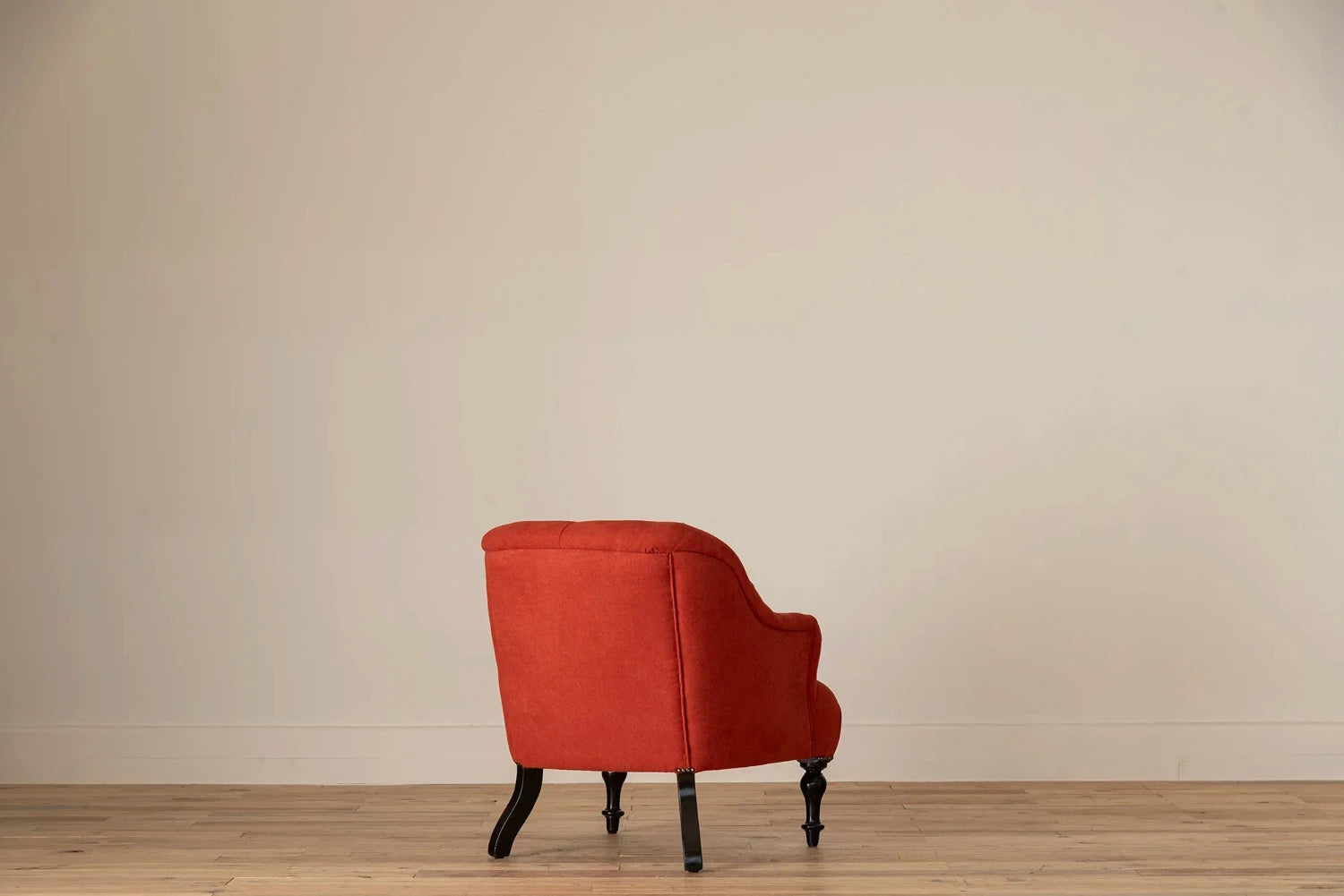 Rust-red button-tufted armchair with nailhead trim and turned dark wood legs, set against a cream wall and light wood floor