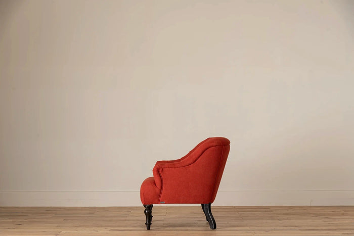 Rust-red button-tufted armchair with nailhead trim and turned dark wood legs, set against a cream wall and light wood floor