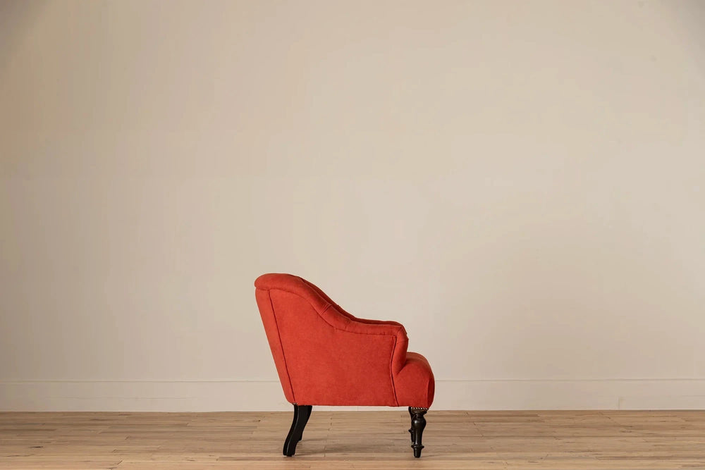 Rust-red button-tufted armchair with nailhead trim and turned dark wood legs, set against a cream wall and light wood floor