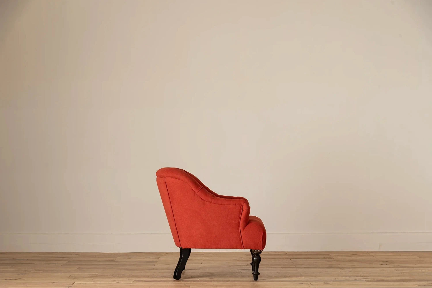 Rust-red button-tufted armchair with nailhead trim and turned dark wood legs, set against a cream wall and light wood floor