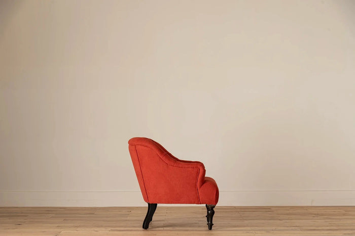 Rust-red button-tufted armchair with nailhead trim and turned dark wood legs, set against a cream wall and light wood floor