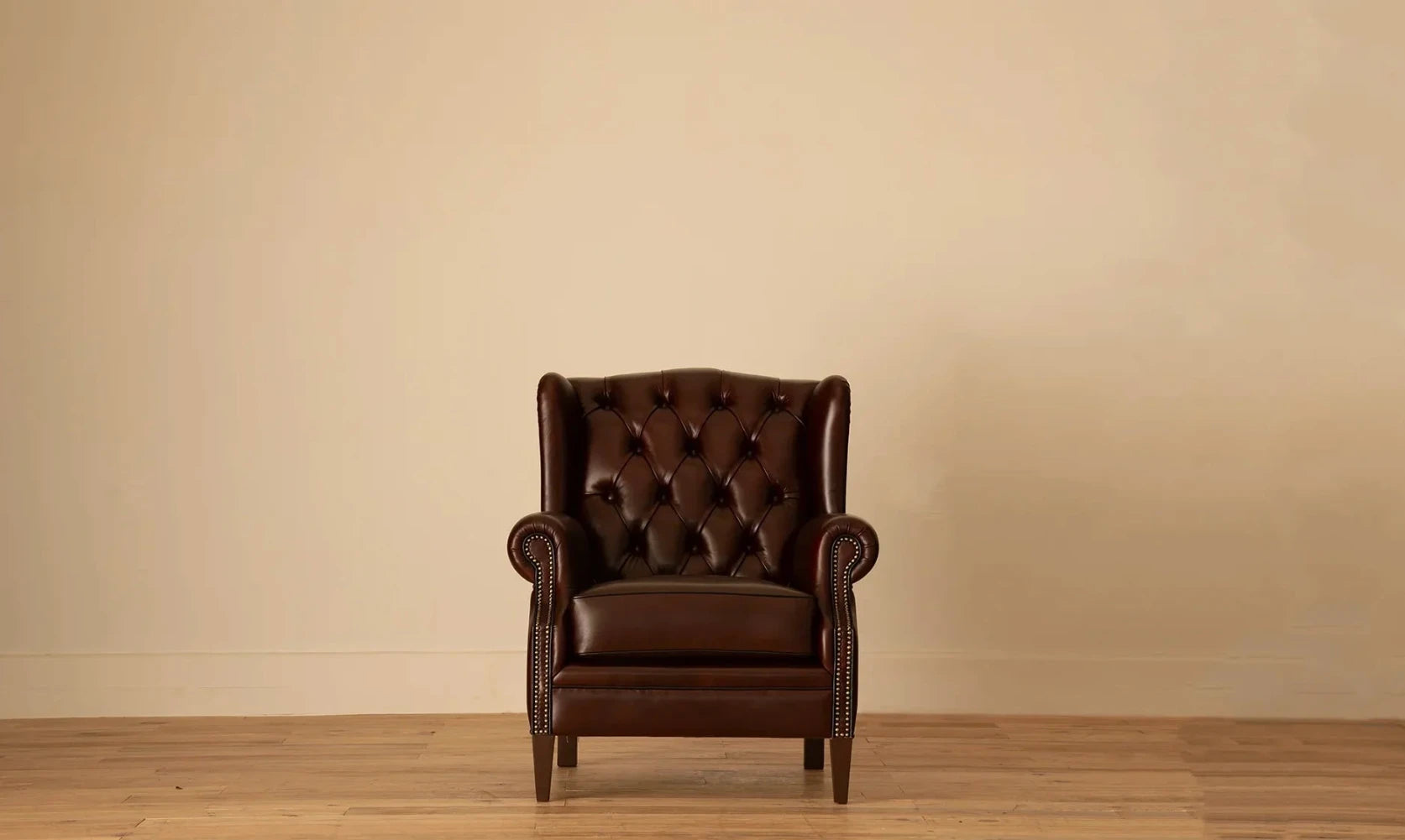 Front view: "James Chesterfield wingback armchair in deep burgundy leather with diamond button tufting and nailhead trim, on tapered wood legs.