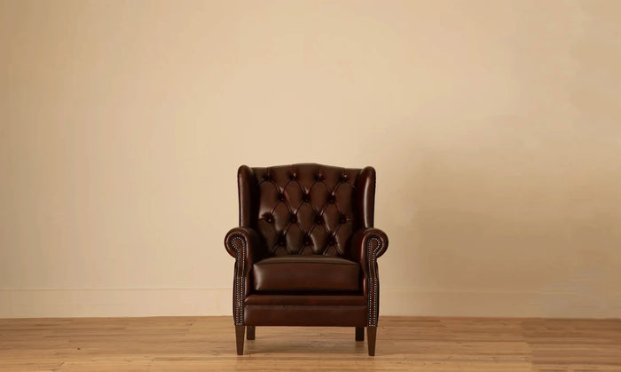 Front view: "James Chesterfield wingback armchair in deep burgundy leather with diamond button tufting and nailhead trim, on tapered wood legs.